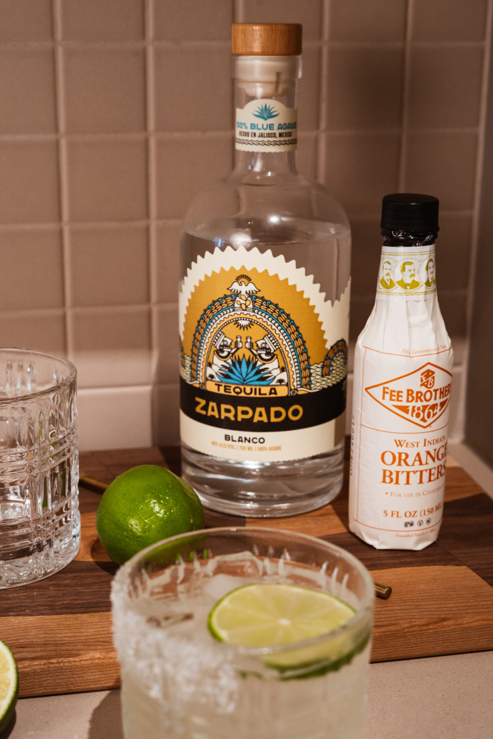 tequila bottle and bitters bottle on wooden cutting board with blurred cocktail in foreground.