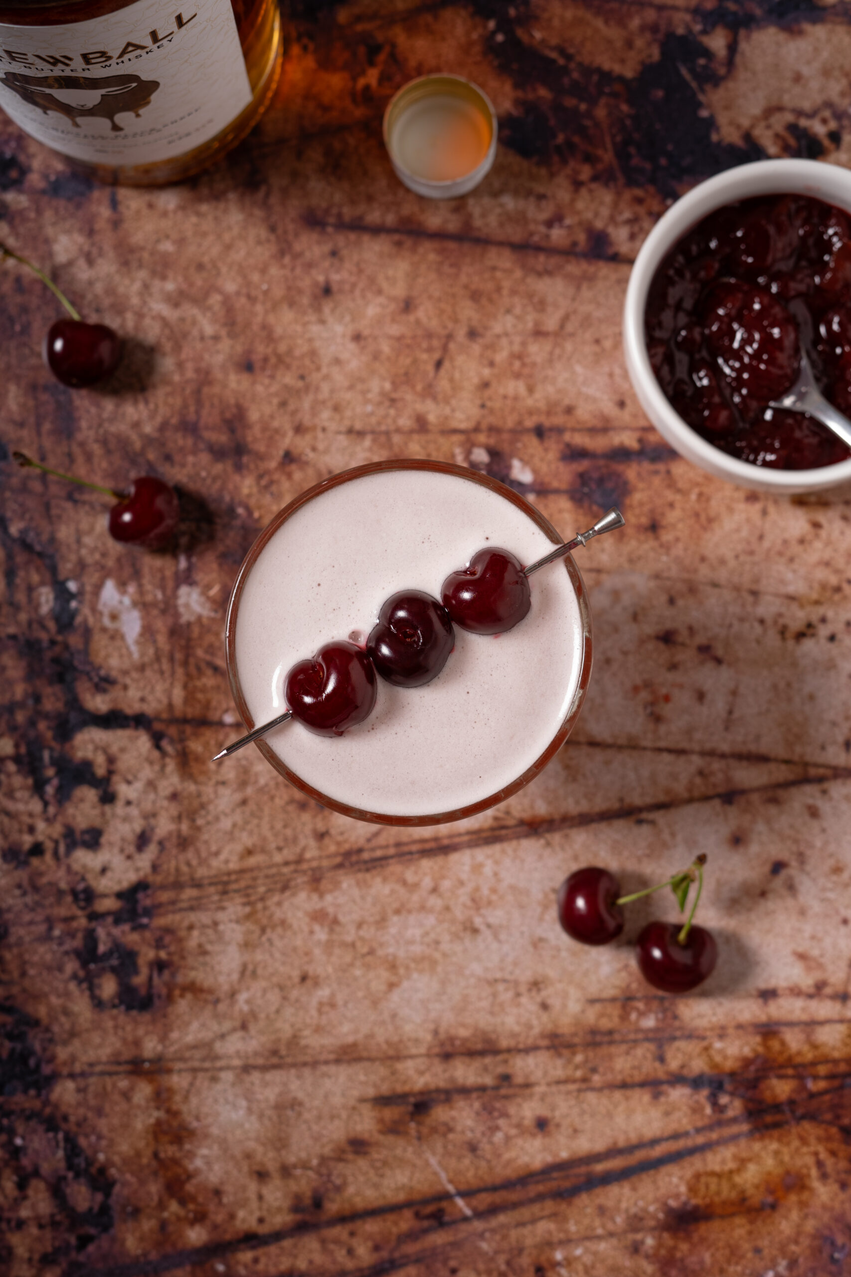 overhead view of whiskey sour with cherry garnish in coupe glass with jam in white bowl.