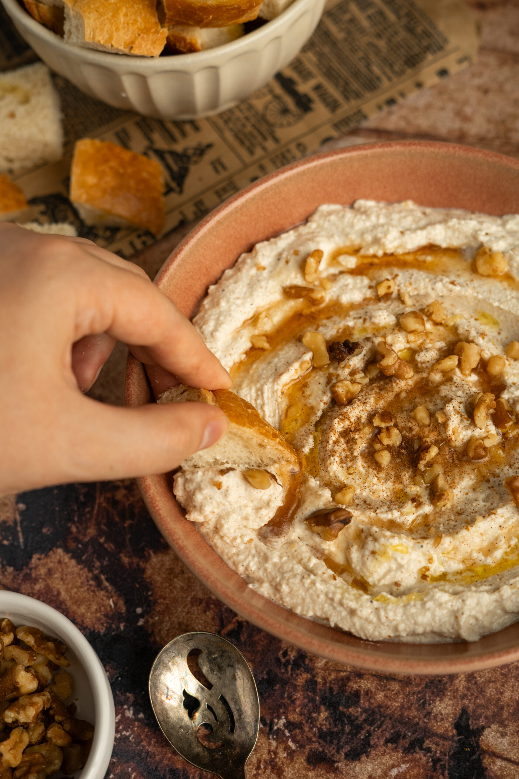 hand dipping bread into whipped feta dip in pink bowl with syrup and walnuts.