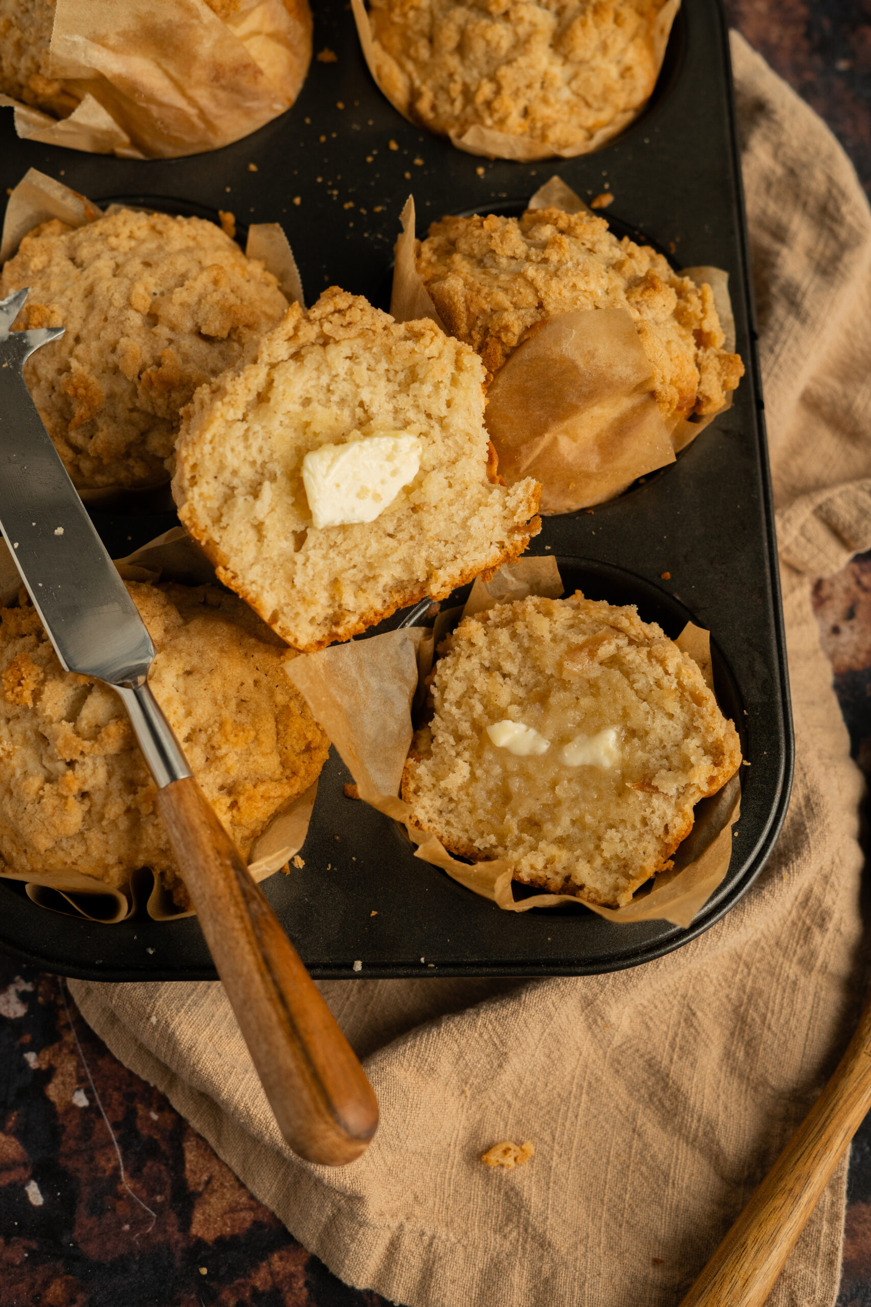 apple muffins in baking tin with one muffin sliced with butter spread on it.