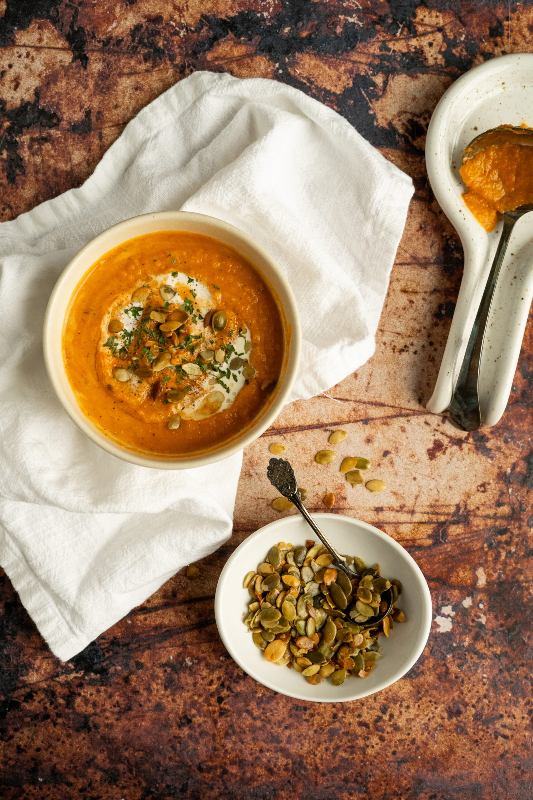 roasted vegetable soup in white bowl with roasted seeds on top, sitting on top of white napkin and brown background.