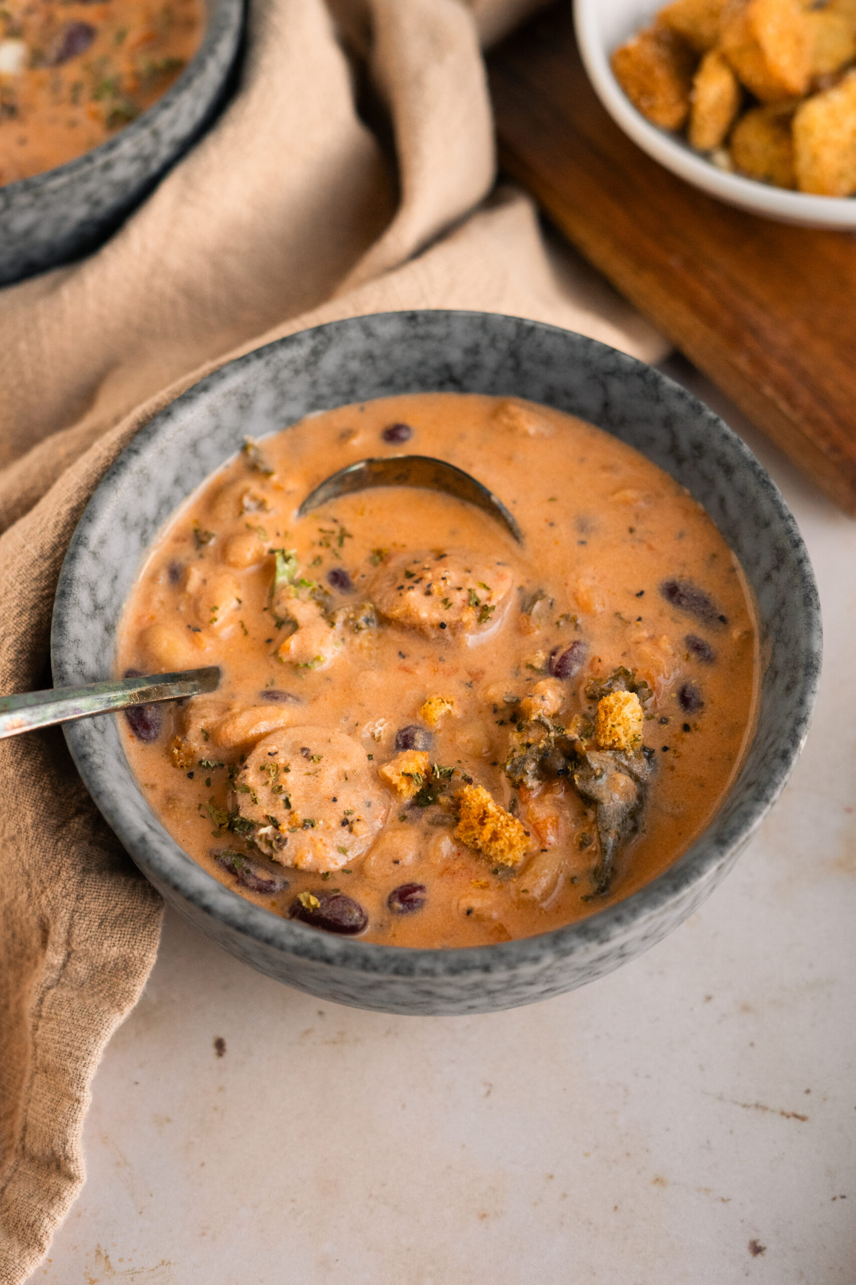 close up of bean and sausage soup in a blue bowl on a beige background.