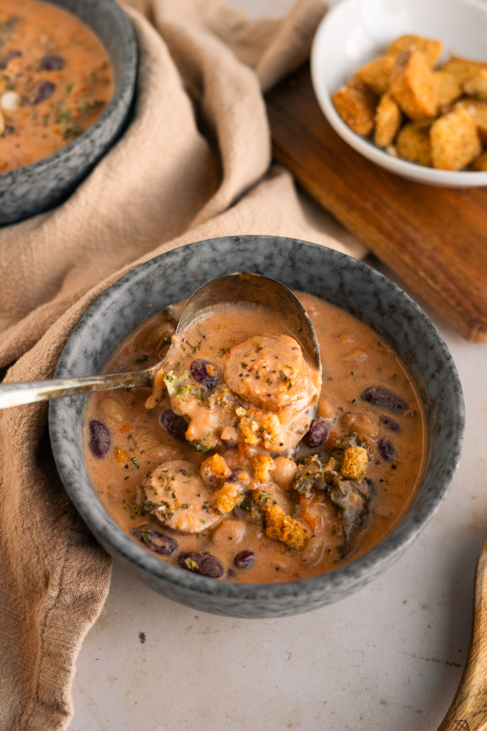 bean and sausage soup in a blue bowl on a beige background with a spoonful of soup over the bowl.