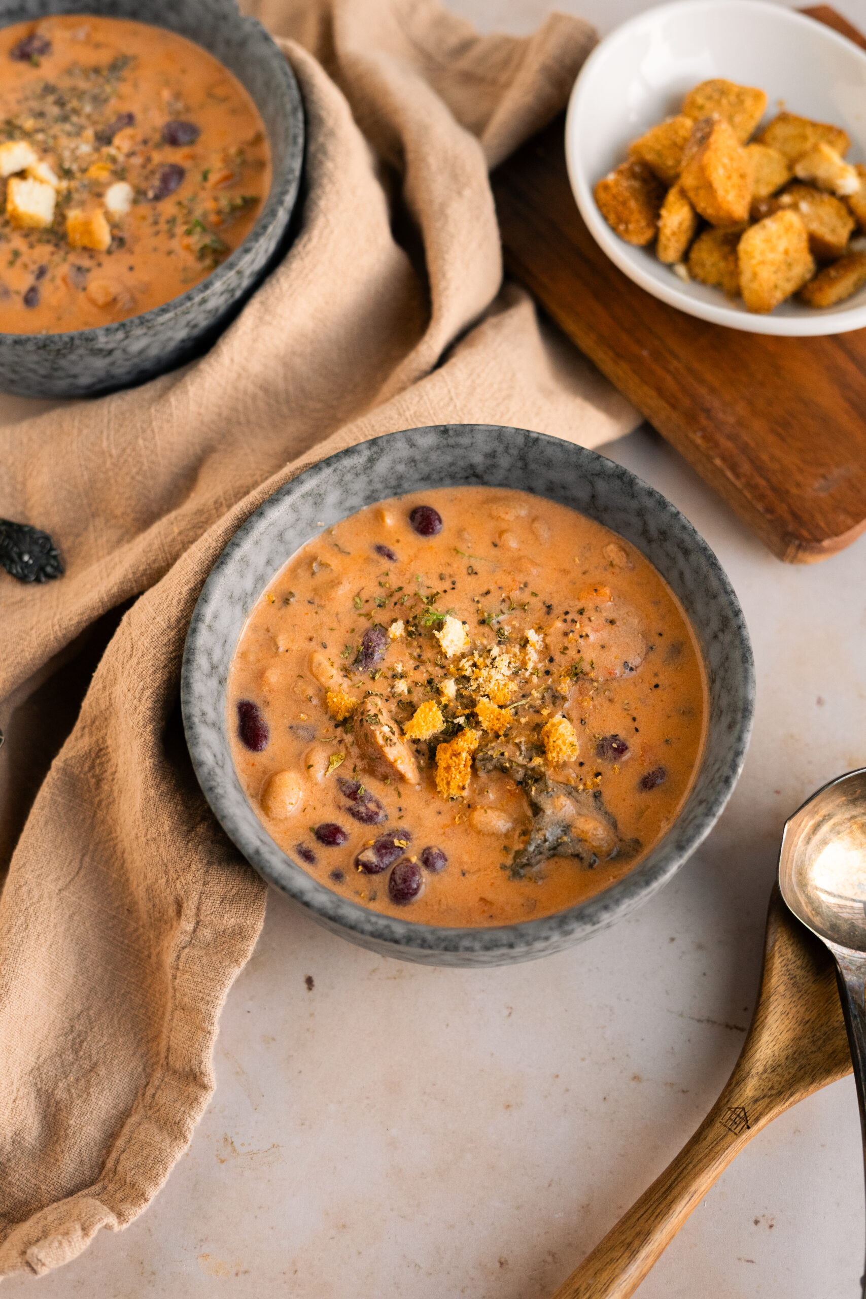 bean and sausage soup in a blue bowl on a beige background with more soup bowls in the back.