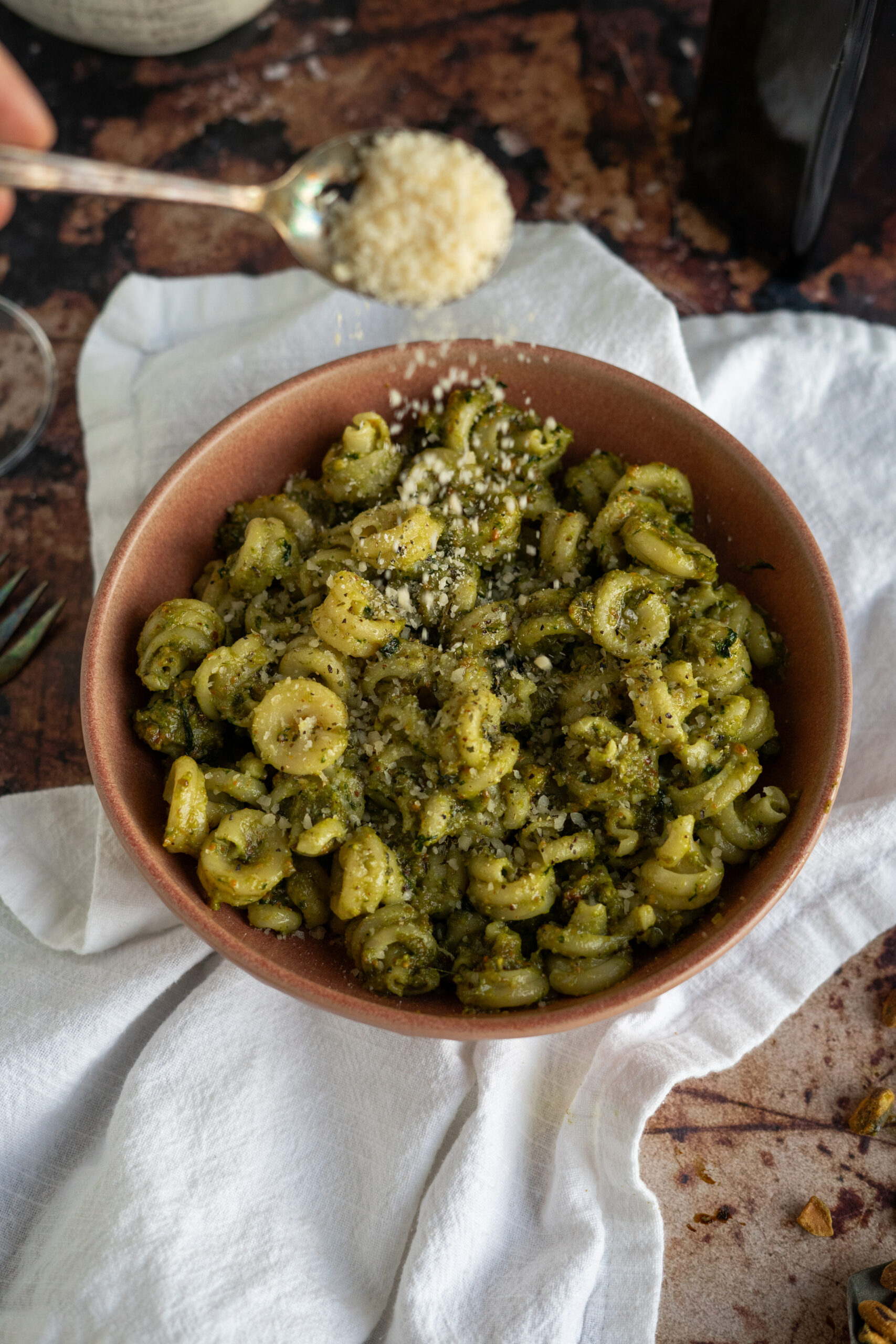 pistachio pesto pasta in pink bowl on white napkin with parmesan cheese sprinkling on top.