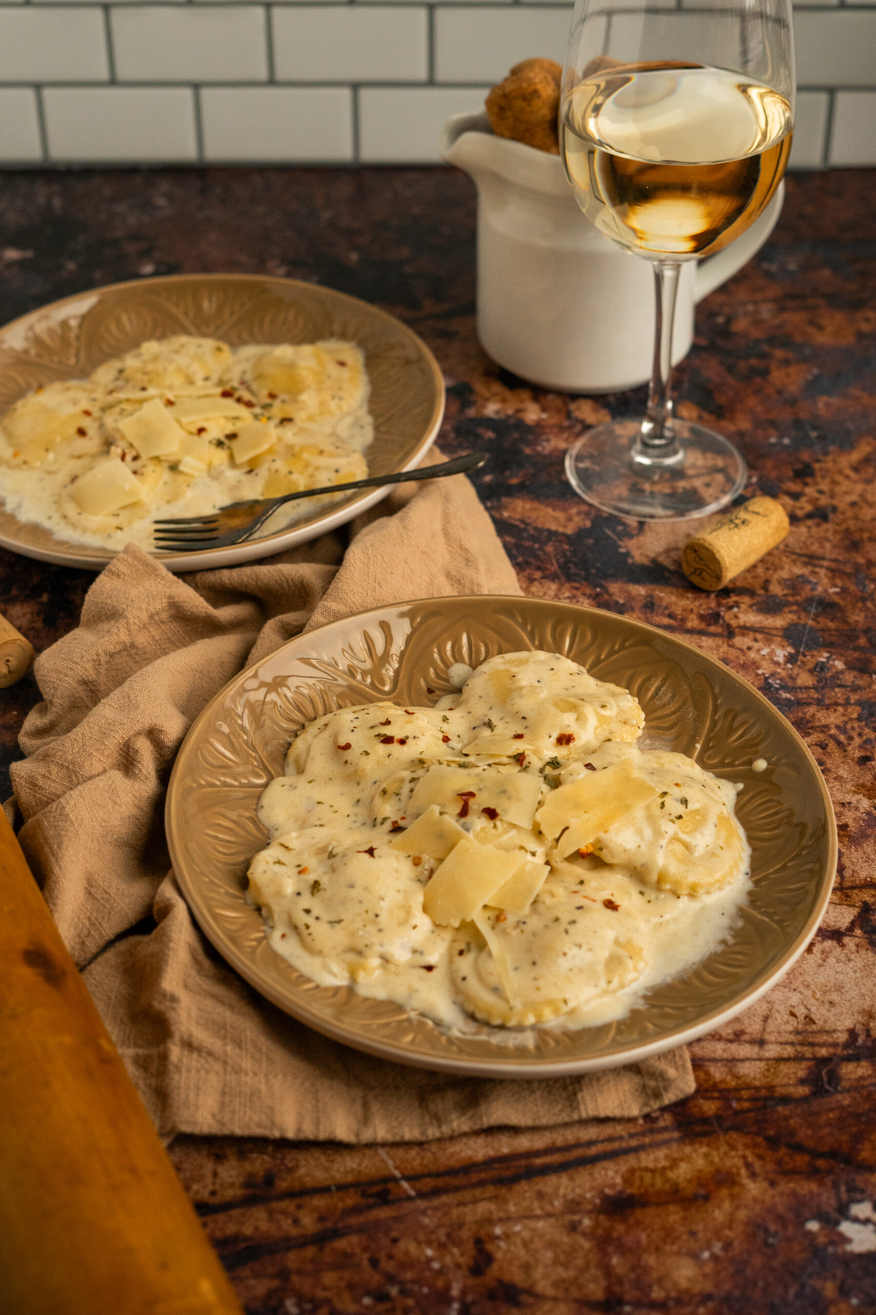 side angle of two plates of ravioli in cream sauce with wine glass behind them.