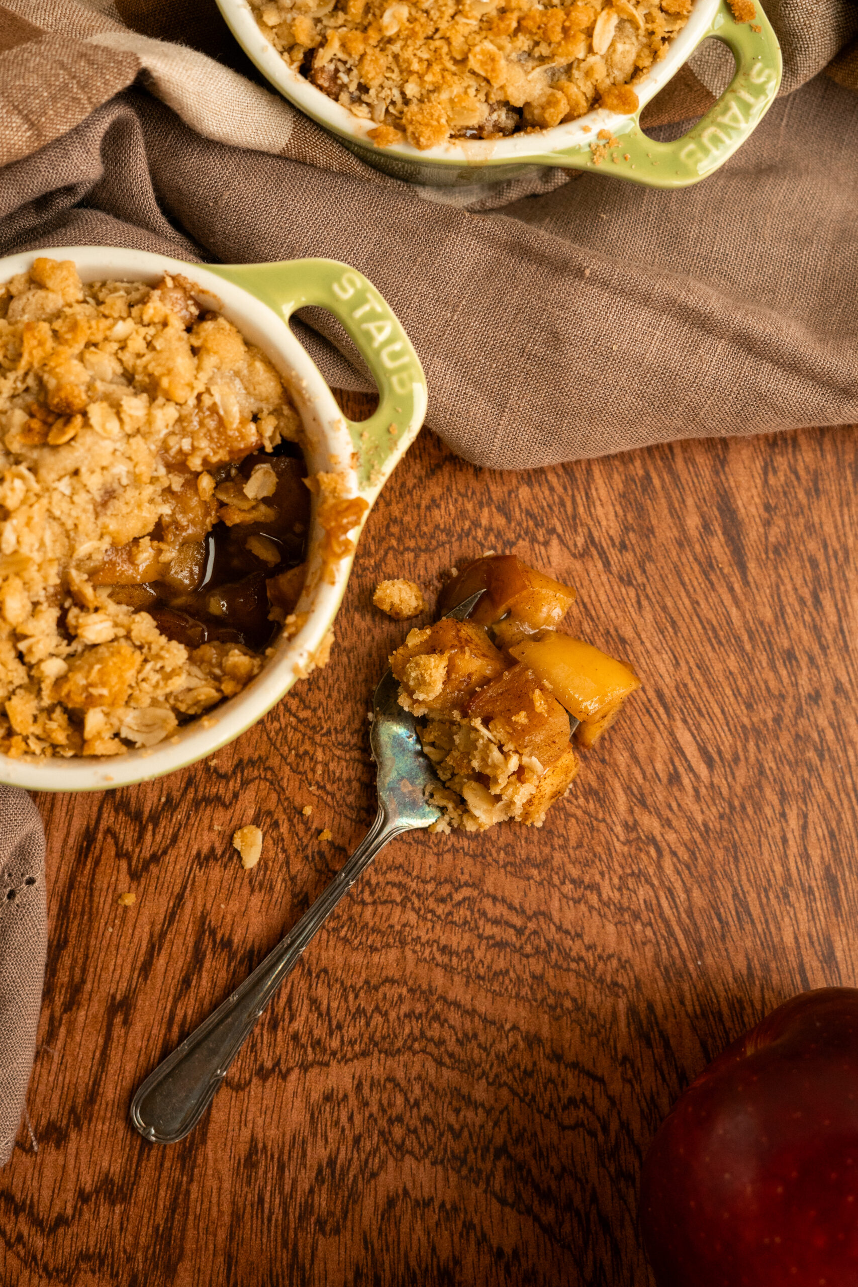 forkful of apple crisp on wooden surface with dish next to fork.