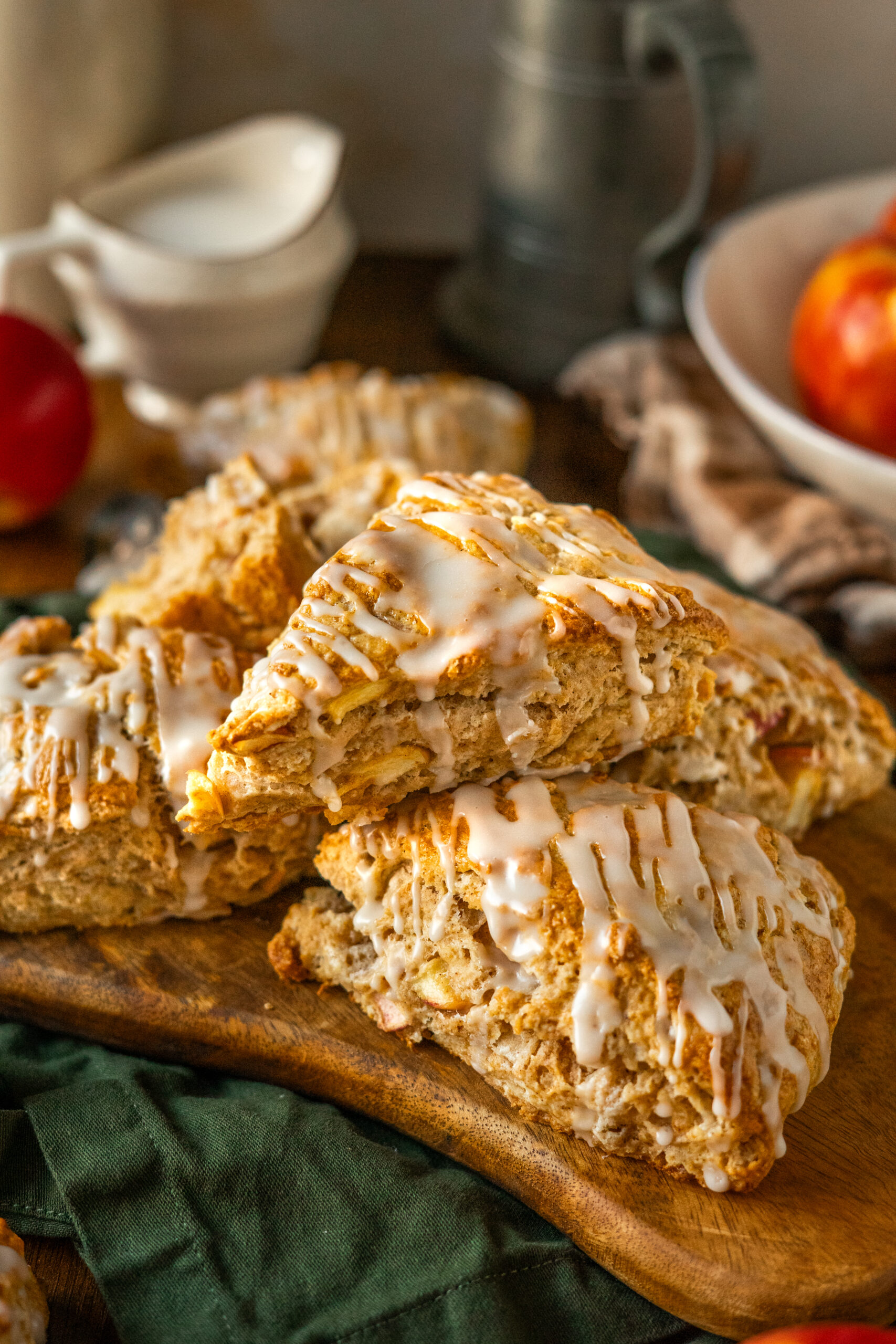 apple cider scones stacked on wooden serving board with apples in bowl in background.