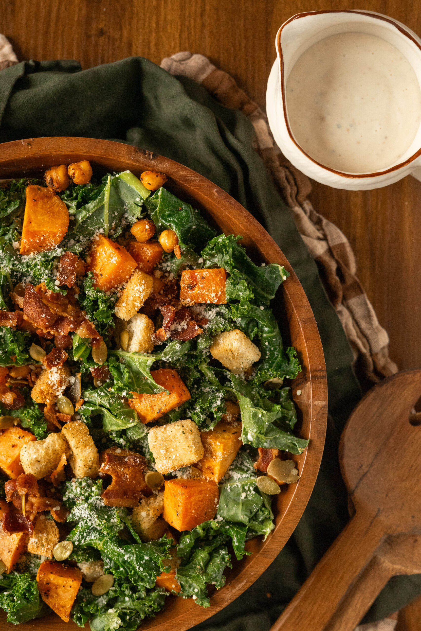 kale caesar salad in wooden bowl with dressing in a pouring dish next to bowl.
