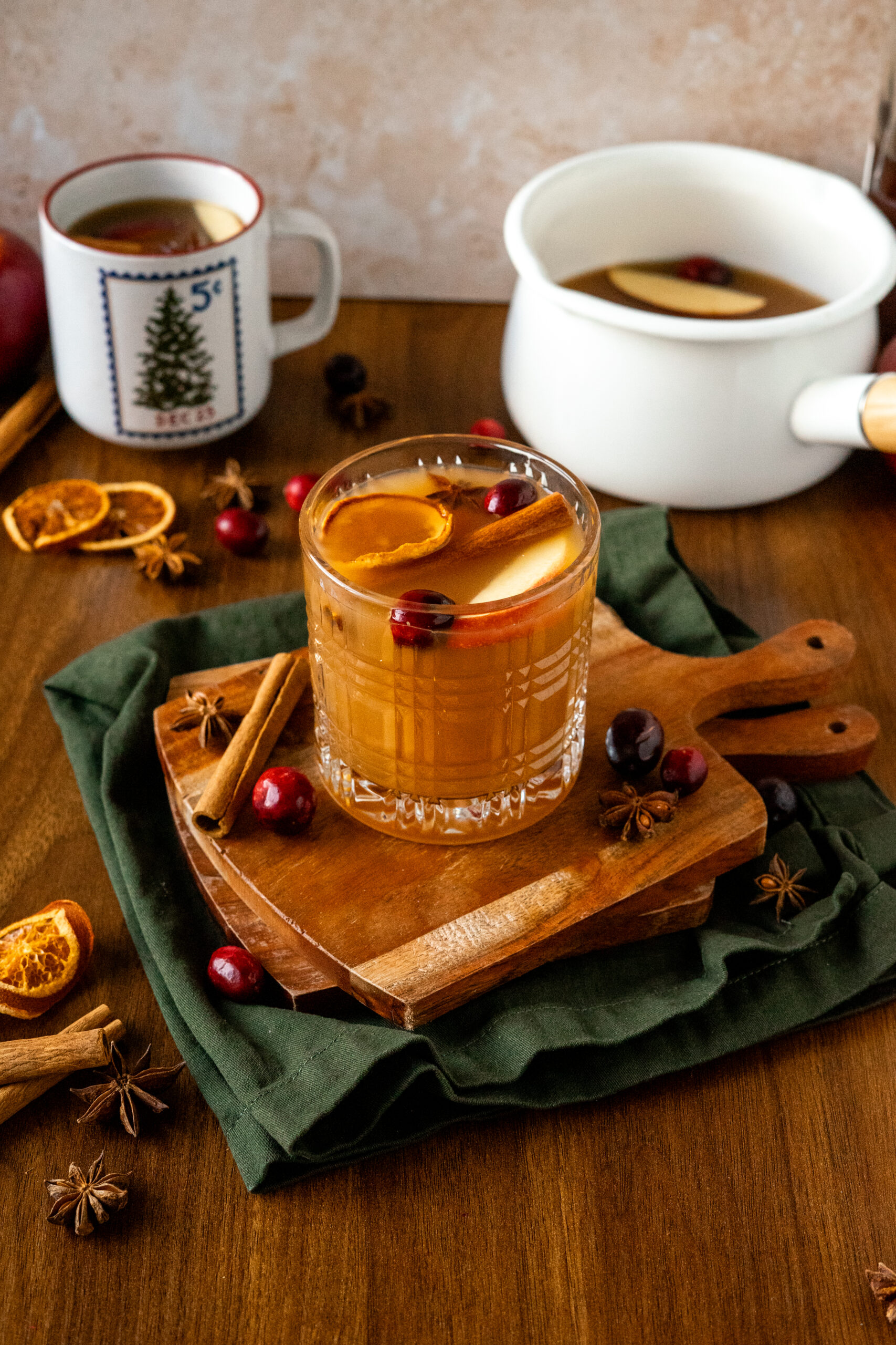 glass of apple cider cocktail on wooden coasters with simmer pot in the background.